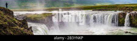 Panoramablick über den mächtigen Wasserfall Godafoss mit einsamem Reisenden, der an seiner Klippe, Island, steht Stockfoto