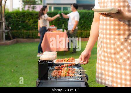 Asiatischer Mann, der grillt und Wurst für eine Gruppe von Freunden kocht, um im Garten zu Hause Party zu essen. Gruppe von Freunden, die Gartengrill la im Freien haben Stockfoto