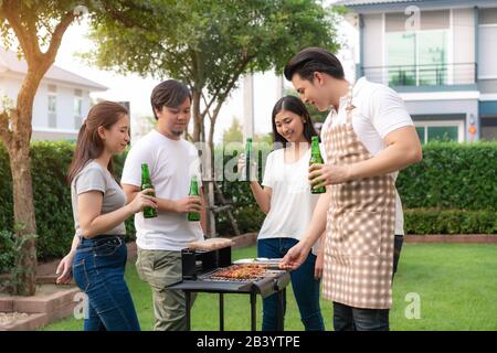 Asiatischer Mann, der grillt und Wurst für eine Gruppe von Freunden kocht, um im Garten zu Hause Party zu essen. Gruppe von Freunden, die Gartengrill la im Freien haben Stockfoto