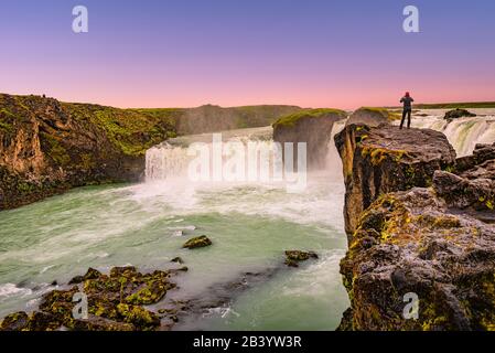 Mächtiger Wasserfall Godafoss bei schönem roten Sonnenuntergang mit einsamem Reisenden, der an seiner Klippe, Island, im Sommer steht Stockfoto