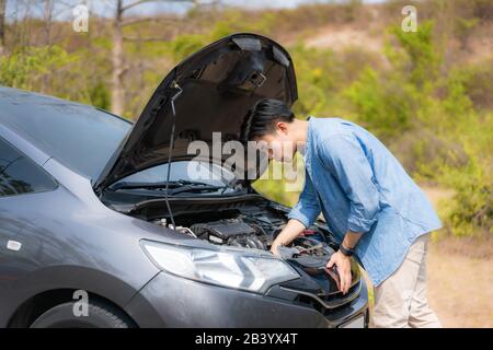 Junger, unzufriedener Asiate, der vor der offenen Haube einen zerbrochenen Automotor Auf der Country Road Inspiziert Und Auf den Straßendienst wartet. Stockfoto