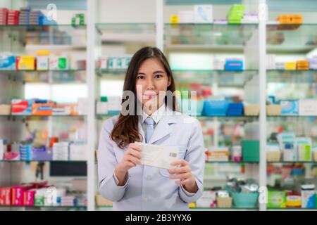 Asiatische junge Frau Apotheker mit einem reizenden Lächeln Holding medicinebox und Kamera in der Apotheke und Drogerie. Medizin, Pharmazie Stockfoto