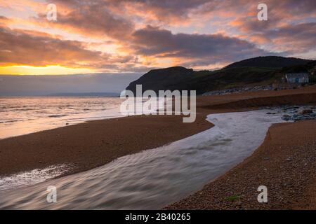 Seatown, Dorset, Großbritannien. März 2020. Wetter in Großbritannien. Die sich abzeichnenden Wolken leuchten bei Sonnenuntergang in Seatown in Dorset nach einem Tag starken Regens mit dem Fluss Winniford, der über den Strand fließt, orange und in der Ferne befindet sich die markante Klippe von Golden Cap, die die höchste Klippe an der Südküste Englands ist. Bildnachweis: Graham Hunt/Alamy Live News Stockfoto