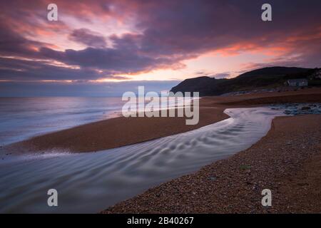 Seatown, Dorset, Großbritannien. März 2020. Wetter in Großbritannien. Die sich abzeichnenden Wolken leuchten bei Sonnenuntergang in Seatown in Dorset nach einem Tag starken Regens mit dem Fluss Winniford, der über den Strand fließt, orange und in der Ferne befindet sich die markante Klippe von Golden Cap, die die höchste Klippe an der Südküste Englands ist. Bildnachweis: Graham Hunt/Alamy Live News Stockfoto