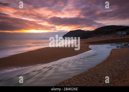 Seatown, Dorset, Großbritannien. März 2020. Wetter in Großbritannien. Die sich abzeichnenden Wolken leuchten bei Sonnenuntergang in Seatown in Dorset nach einem Tag starken Regens mit dem Fluss Winniford, der über den Strand fließt, orange und in der Ferne befindet sich die markante Klippe von Golden Cap, die die höchste Klippe an der Südküste Englands ist. Bildnachweis: Graham Hunt/Alamy Live News Stockfoto