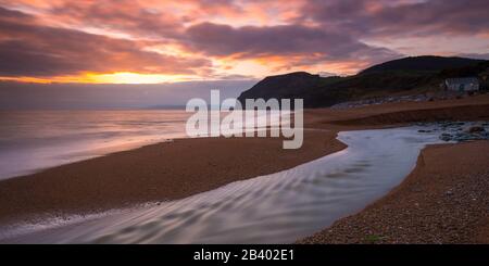 Seatown, Dorset, Großbritannien. März 2020. Wetter in Großbritannien. Die sich abzeichnenden Wolken leuchten bei Sonnenuntergang in Seatown in Dorset nach einem Tag starken Regens mit dem Fluss Winniford, der über den Strand fließt, orange und in der Ferne befindet sich die markante Klippe von Golden Cap, die die höchste Klippe an der Südküste Englands ist. Bildnachweis: Graham Hunt/Alamy Live News Stockfoto