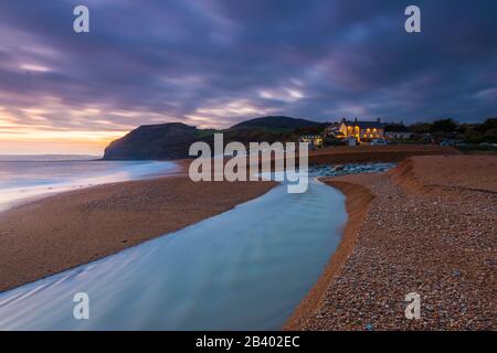 Seatown, Dorset, Großbritannien. März 2020. Wetter in Großbritannien. Blick auf den Strand in Seatown in Dorset kurz nach Sonnenuntergang nach einem Tag starken Regens mit dem Fluss Winniford, der über den Strand fließt, und in der Ferne befindet sich die markante Klippe von Golden Cap, die die höchste Klippe an der Südküste Englands ist. Neben dem Strand befindet sich das Anchor Inn. Bildnachweis: Graham Hunt/Alamy Live News Stockfoto