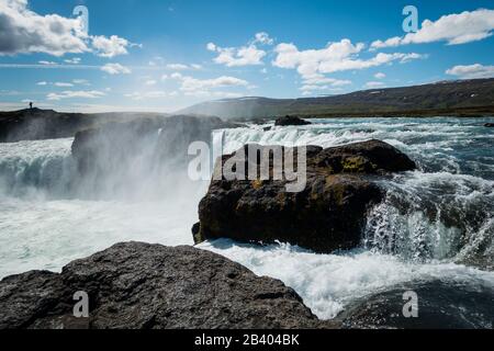 Mächtiger Wasserfall von Godafoss in der Nähe von Akureyri Island an einem schönen sonnigen Tag mit Nebel, der in der Luft hängt. Blauer Himmel mit ein paar Wolken. Silhouette einer Person, die in der Ferne steht. Stockfoto