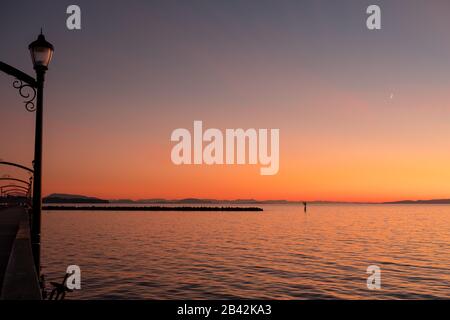 Der Himmel von Orangen, Rottönen, Pinks und Gelbtönen spiegelt sich in ruhigen Gewässern der Semiahmoo Bay, White Rock, BC, Kanada, wider. Oben schwebt ein Neumond in den Himmel. Stockfoto