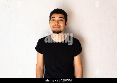 Junger hispanischer Mann mit schwarzer Kleidung, die mit einem einfachen Lächeln auf weißem Hintergrund posiert Stockfoto