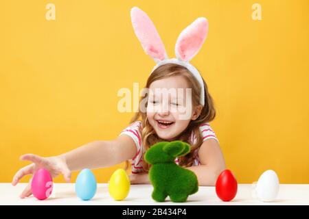Osterkonzept. Glückliches Kind in den Hünenohren gelber Hintergrund. Ein kleines Mädchen sitzt an einem Tisch und jagt nach farbigen Eiern. Stockfoto