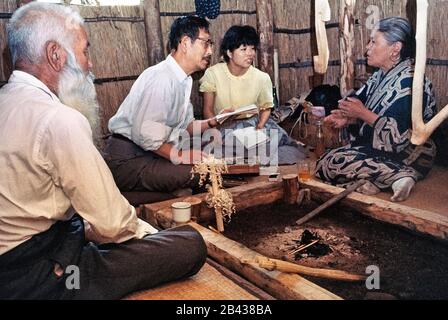 Der japanische Sprachwissenschaftler Professor Kyosuke Kindaichi und sein weibliches Assistenteninterview eine ältere Ainu-Frau in traditionellem Kleid, als ihr bärtiger Ehemann ihre Geschichten in ihrer Heimat auf der Insel Hokkaido im Norden Japans hört. Sie waren zwei von nur 300 reinblütigen Ainu (ausgesprochen i-noo), die noch lebten, als diese historische Fotografie 1962 aufgenommen wurde. Seit dieser Zeit haben sich die Ainu in die japanische Gesellschaft eingenistet und ihre uralte Lebensweise wird heute nur noch in besonderen Touristendörfern erblickt. Die Ainu wurden 2008 offiziell als Ureinwohner Japans anerkannt. Stockfoto