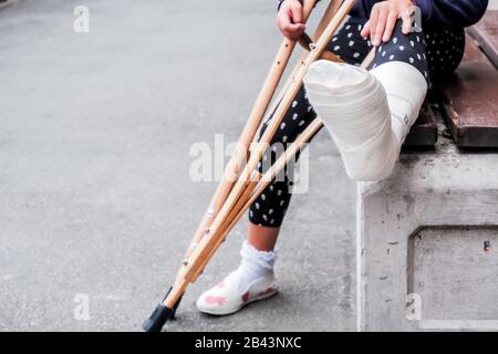 Unkenntlich Mädchen sitzt auf der Straße auf einer Bank mit Ein gebrochenes Bein und Krücken.Behinderte Mädchen mit Krücken.ein Unfall während Springen auf einem Trampolin Stockfoto