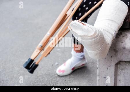 Unkenntlich Mädchen sitzt auf der Straße auf einer Bank mit Ein gebrochenes Bein und Krücken.Behinderte Mädchen mit Krücken.ein Unfall während Springen auf einem Trampolin Stockfoto