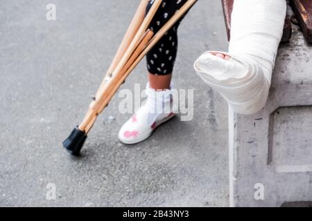 Kind mit Krücken und gebrochenen Beinen für das Gehen, ein Unfall bei Sprüngen. Gebrochenes Bein, Holzkrücken, Kind mit gebrochenem Bein in weißem Gips gegossen Stockfoto