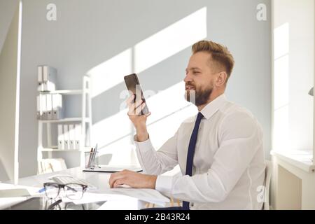 Ein Unternehmer, der mit viel beschäftigt ist, liest die Nachricht am Telefon an einem Tisch mit einem Computer im Büro. Stockfoto