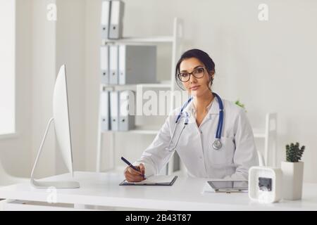 Selbstbewusste Frau Ärztin Kinderärztin schreibt in einer Zwischenablage, die an einem Tisch in einem weißen Büro des Krankenhauses sitzt Stockfoto