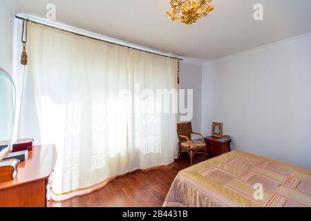 Ein großes Schlafzimmer mit Mahagoni, Korbstühlen und einem eleganten Schminktisch mit einem halbkreisförmigen Spiegel. Das Bett hat eine beige Decke in Quadraten. Stockfoto