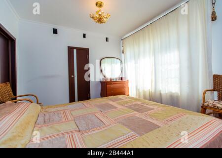 Ein großes Schlafzimmer mit Mahagoni, Korbstühlen und einem eleganten Schminktisch mit einem halbkreisförmigen Spiegel. Das Bett hat eine beige Decke in Quadraten. Stockfoto