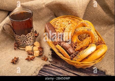 Plätzchen, Bagels, Schokoladenkuchen und Sternanis in einem Korbkorb und ein Glas Tee in einem Vintage-Cup-Halter auf dem Hintergrund rauer Homespun-Qualität Stockfoto