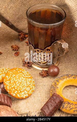 Ein Glas Tee in einem Vintage-Cup-Halter und Plätzchen, Bagels, Schokolade, Sternanis auf dem Hintergrund rauer Homespun-Qualität. Nahaufnahme Stockfoto