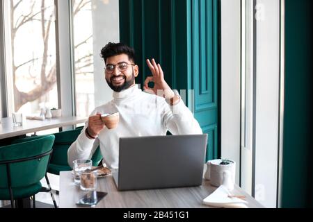 Gutaussehender Geschäftsmann, der im Café sitzt und arbeitet. Es gibt einen Laptop, ein Smartphone und eine Tasse Kaffee. Mann zeigt Geste OK und lächelt. Stockfoto