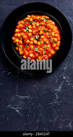 Punjabi Chole Masala oder Chana Masala mit Garam Masala-Gewürzen, Tomatensauce, Lorbeerblatt, mit Petersilie auf einem schwarzen Teller bestreut, auf einer dunklen Betonba Stockfoto