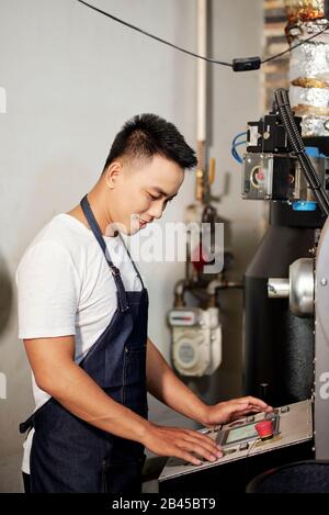 Lächelnder vietnamesischer junger Mann mit Denimschürze beim Betrieb einer neuen Kaffeeröstmaschine Stockfoto