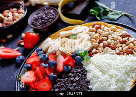 Süße, leuchtende asiatische Mondpfannkuchen mit Schokoladenchips, Erdnüssen, Erdbeeren, Blaubeeren, Banane, Käse, auf dunklem Betongrund mit Zutaten Stockfoto