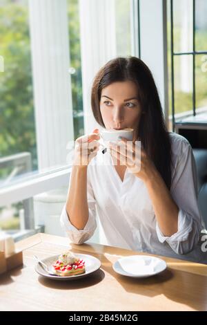 Schönen lächelnden jungen Frau im Cafe in der Nähe der Fenster mit Kaffee und leckeren Kuchen. Stockfoto