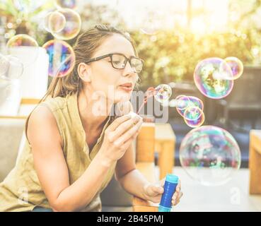 Junge gutaussehende Frau, die Seifenblase in der Stadtbar mit hinterem Sonnenschein bläst - Porträt des glücklichen Mädchens, das im Freien Spaß hat - Glücklichsein und Freiheitskonzept - Stockfoto