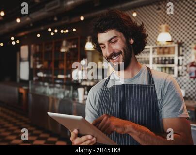 Fröhlicher junger kaukasischer Café-Besitzer, der Schürze mit digitalem Tablet trägt Stockfoto