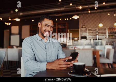 Fröhlicher, gutaussehender junger afrikanischer Mann, der sich im modernen Café mit dem Handy beim Kaffeetrinken entspannen kann