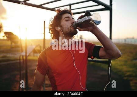 Nahaufnahme eines kaukasischen jungen Mannes mit Ohrhörern im Ohr Trinkwasser nach dem Training im Sportpark im Freien Stockfoto