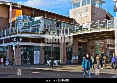 Die Menschen schlendern im Winter unter der Sonne unter den vielen Bars, Geschäften und Restaurants im Mermaid Quay in Cardiff Bay. Die Gegend ist bekannt als "Tacoma-Platz". Stockfoto