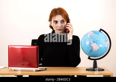 Junge Reisebüro-Frau hinter dem Schreibtisch mit Laptop und geografischer Weltöffentlichkeit, die auf einem Handy spricht. Stockfoto