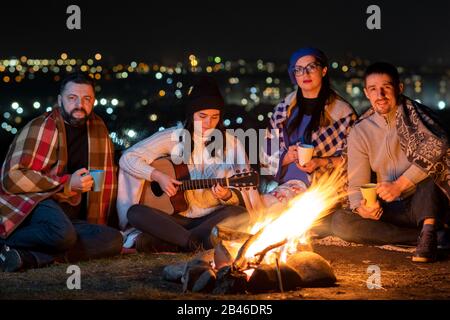 Eine Gruppe von Leuten, die nachts in der Nähe von Lagerfeuer sitzen und Gitarre spielen, Lieder singen und glücklich miteinander sprechen. Stockfoto