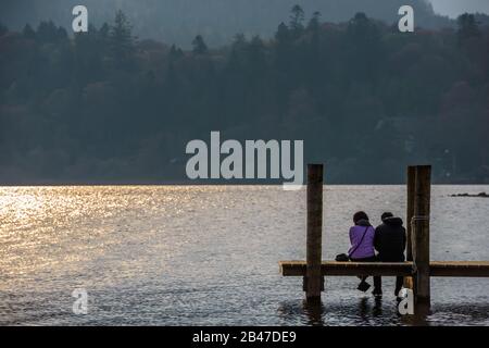 Ein Paar sitzt auf einem Steg und betrachtet den Sonnenuntergang über Derwentwater in Keswick, Lake District National Park, England. Stockfoto