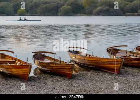 Zwei Männer rudern ein Boot an einer Reihe von hölzernen Ruderbooten am Nordufer von Derwentwater in Keswick im Englischen Lake District bei Sonnenuntergang. Stockfoto