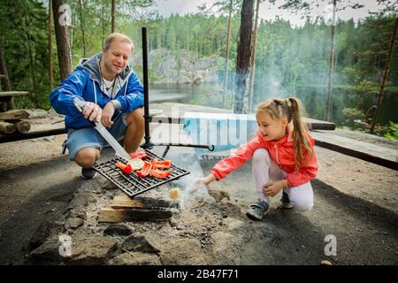 Der Mann und seine kleine Tochter grillen im Wald am felsigen Ufer des Sees und machen ein Feuer, grillen Brot, Gemüse und Marshmallow. Frohes Familienfest Stockfoto