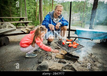 Der Mann und seine kleine Tochter grillen im Wald am felsigen Ufer des Sees und machen ein Feuer, grillen Brot, Gemüse und Marshmallow. Frohes Familienfest Stockfoto