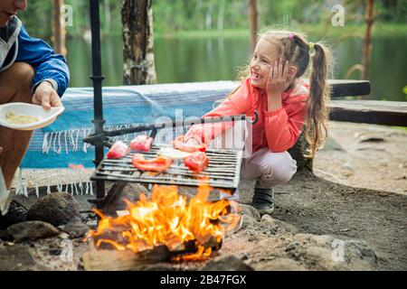 Der Mann und seine kleine Tochter grillen im Wald am felsigen Ufer des Sees und machen ein Feuer, grillen Brot, Gemüse und Marshmallow. Frohes Familienfest Stockfoto