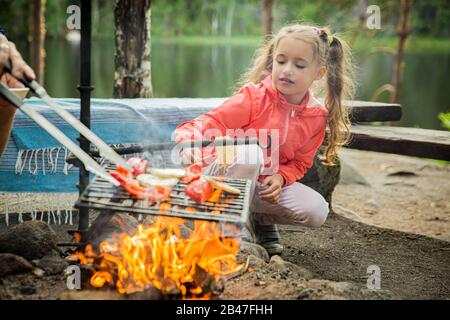 Der Mann und seine kleine Tochter grillen im Wald am felsigen Ufer des Sees und machen ein Feuer, grillen Brot, Gemüse und Marshmallow. Frohes Familienfest Stockfoto