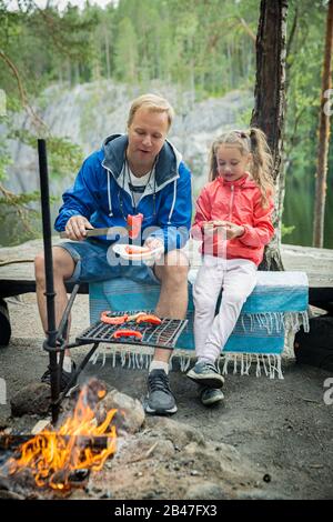 Der Mann und seine kleine Tochter grillen im Wald am felsigen Ufer des Sees und machen ein Feuer, grillen Brot, Gemüse und Marshmallow. Frohes Familienfest Stockfoto