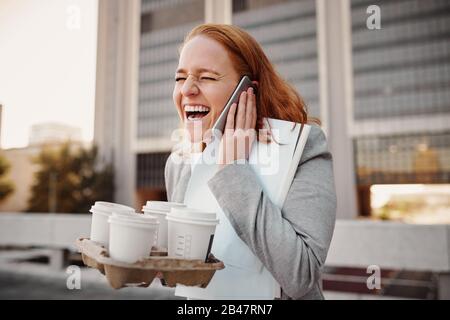 Junge Frau, die Akte mit Dokumenten und Kaffeetablett hält, spricht über Smartphone lacht Stockfoto