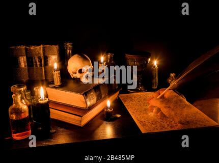 Assistentendesks. Schreibtisch mit Kerzenlicht. Menschlicher Schädel, alte Bücher, Goblet und Tränenflaschen. Gespenstische Hand schreibt auf Pergament mit Federstift. Stockfoto