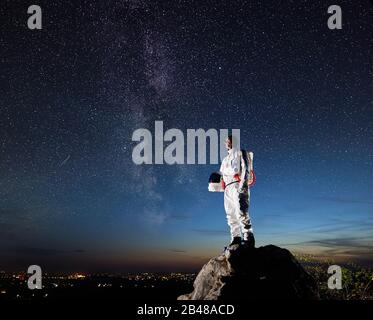 Seitenansicht des Weltraumreisenden, der auf einem felsigen Hügel steht und einen schönen Himmel mit Sternen betrachtet. Kosmonaut im weißen Raumanzug mit Helm. Konzept der Raumfahrt und Kosmonautik. Stockfoto