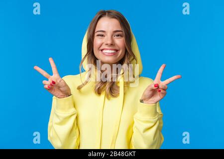 Nahaufnahme Portrait niedlich albern blonde europäische Frau in gelber Kapuze, zeigt Frieden Geste und lächelnd optimistisch, senden positive Stimmung, blau stehend Stockfoto