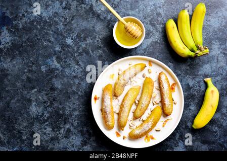 Frittierte Bananen auf weißem Teller über blauem Steinhintergrund mit freiem Textbereich. Leckeres Dessert aus panfrittierten Bananen im asiatischen Stil. Draufsicht, flach Stockfoto