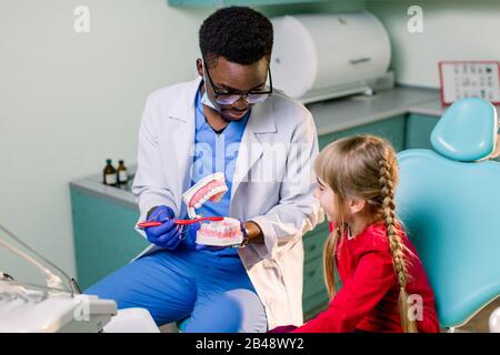 Fröhlicher lächelnder schwarzer Zahnarzt erzählt kaukasischen kleinen Mädchen, wie sie ihre Zähne putzen. Karies Prävention, Zahnheilkunde, Zahnhygiene Konzept. Stockfoto